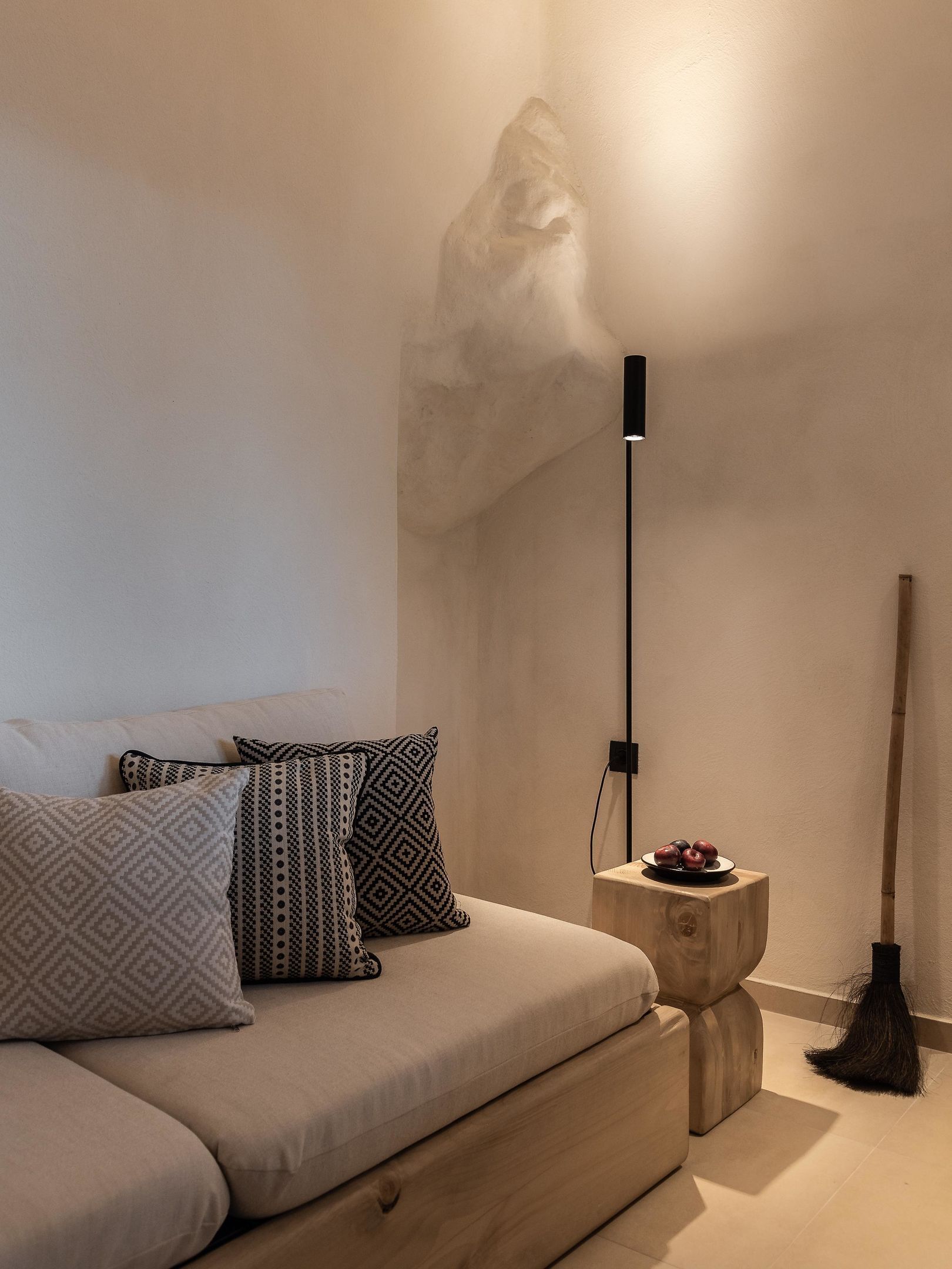 A minimalist living room with a sofa, patterned cushions, a standing lamp, a small wooden side table with fruit, and a broom.
