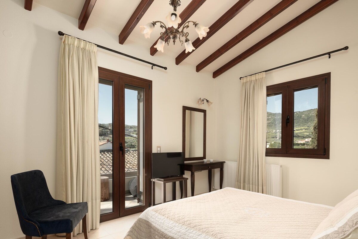 A bedroom with a bed, a chair, a desk, a mirror, and a view of the outdoors.