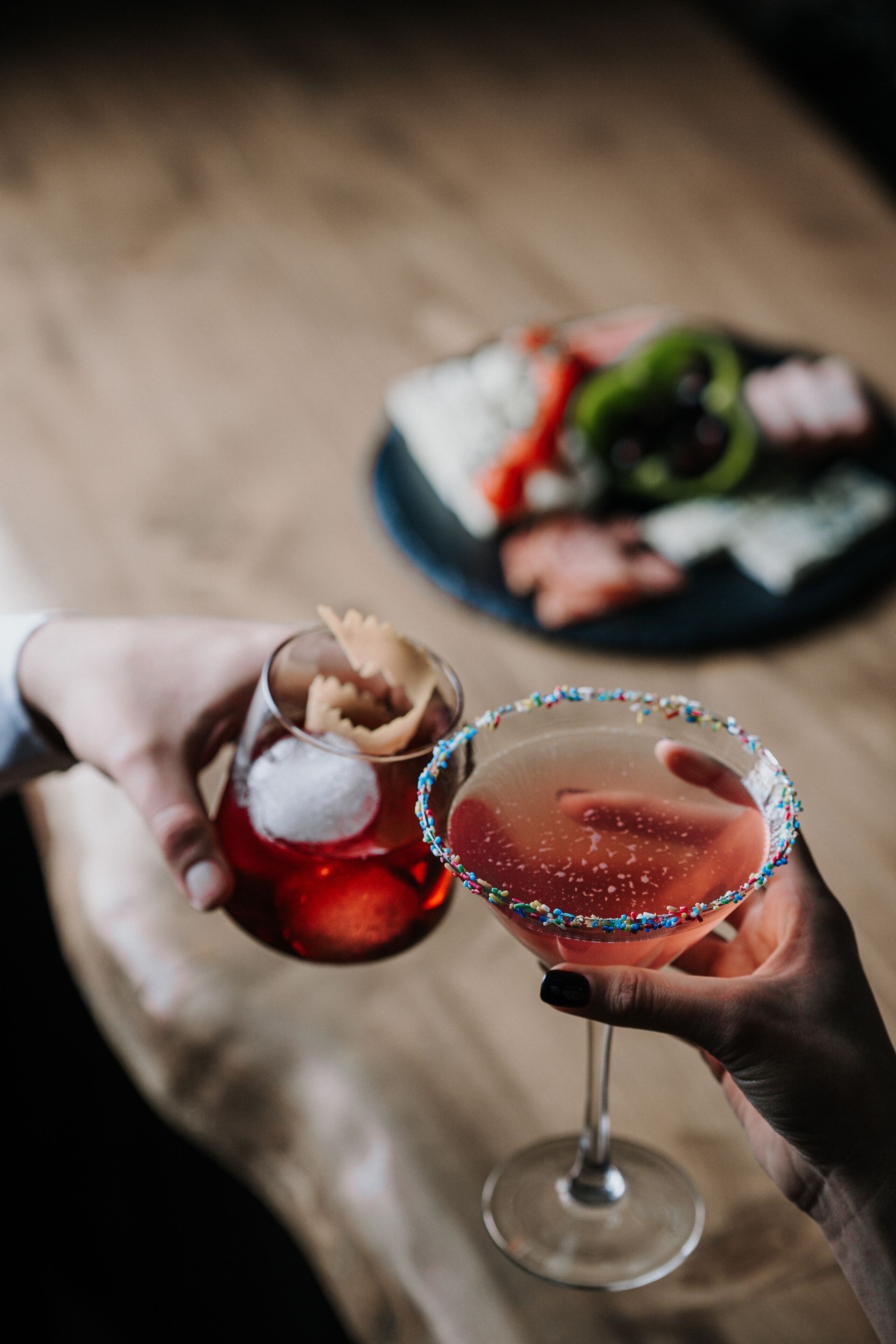 Two hands holding a martini glass and a cocktail glass with a colorful rim.
