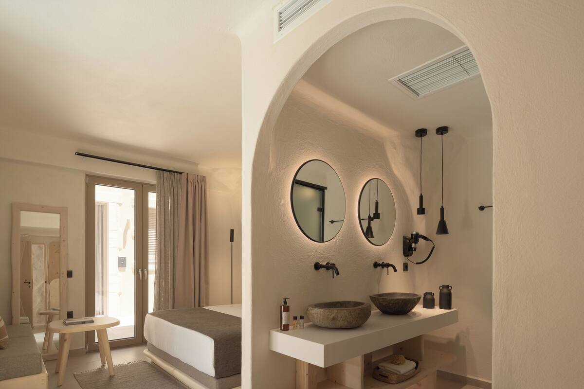 A modern bedroom with a large bed, a small round table, and a bathroom with a large mirror, sink, and towel rack.