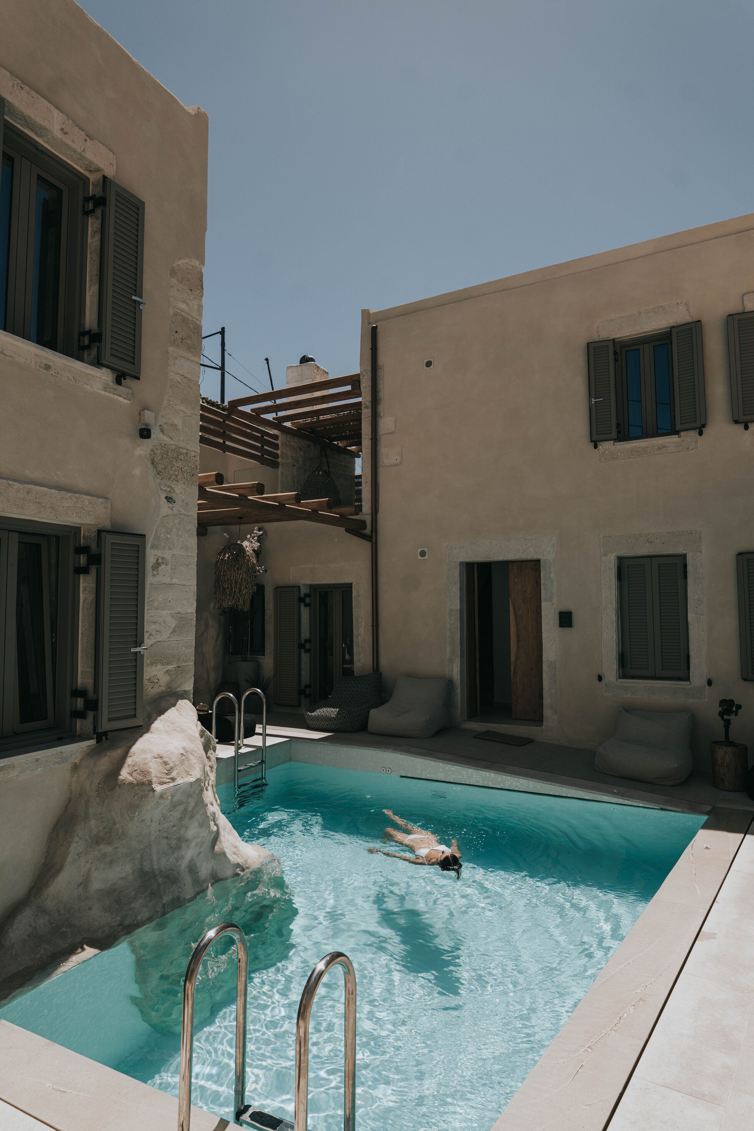 Une piscine où une personne nage, entourée de bâtiments aux murs de pierre et aux balcons en bois.