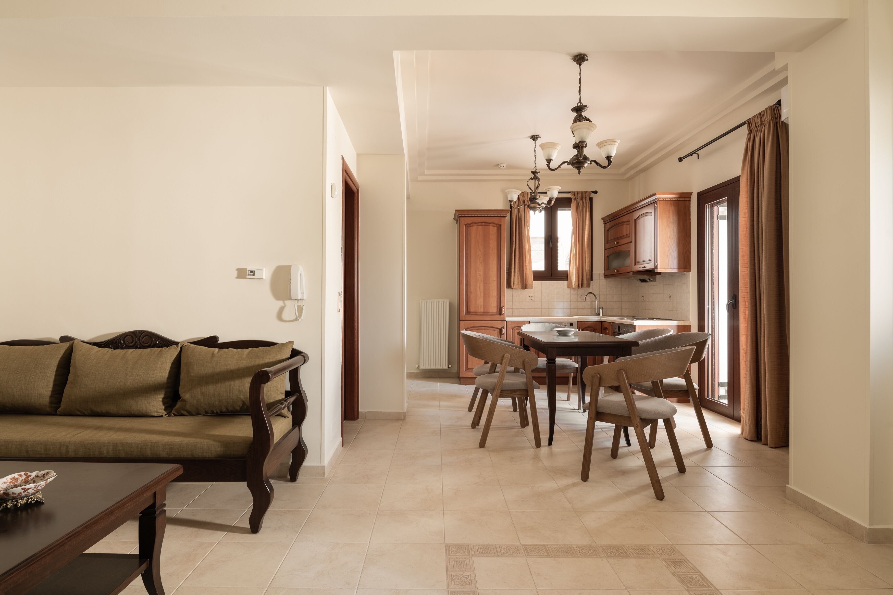 Una sala de estar con un sofá y una mesa de comedor con sillas. Hay una zona de cocina con armarios de madera y una araña.