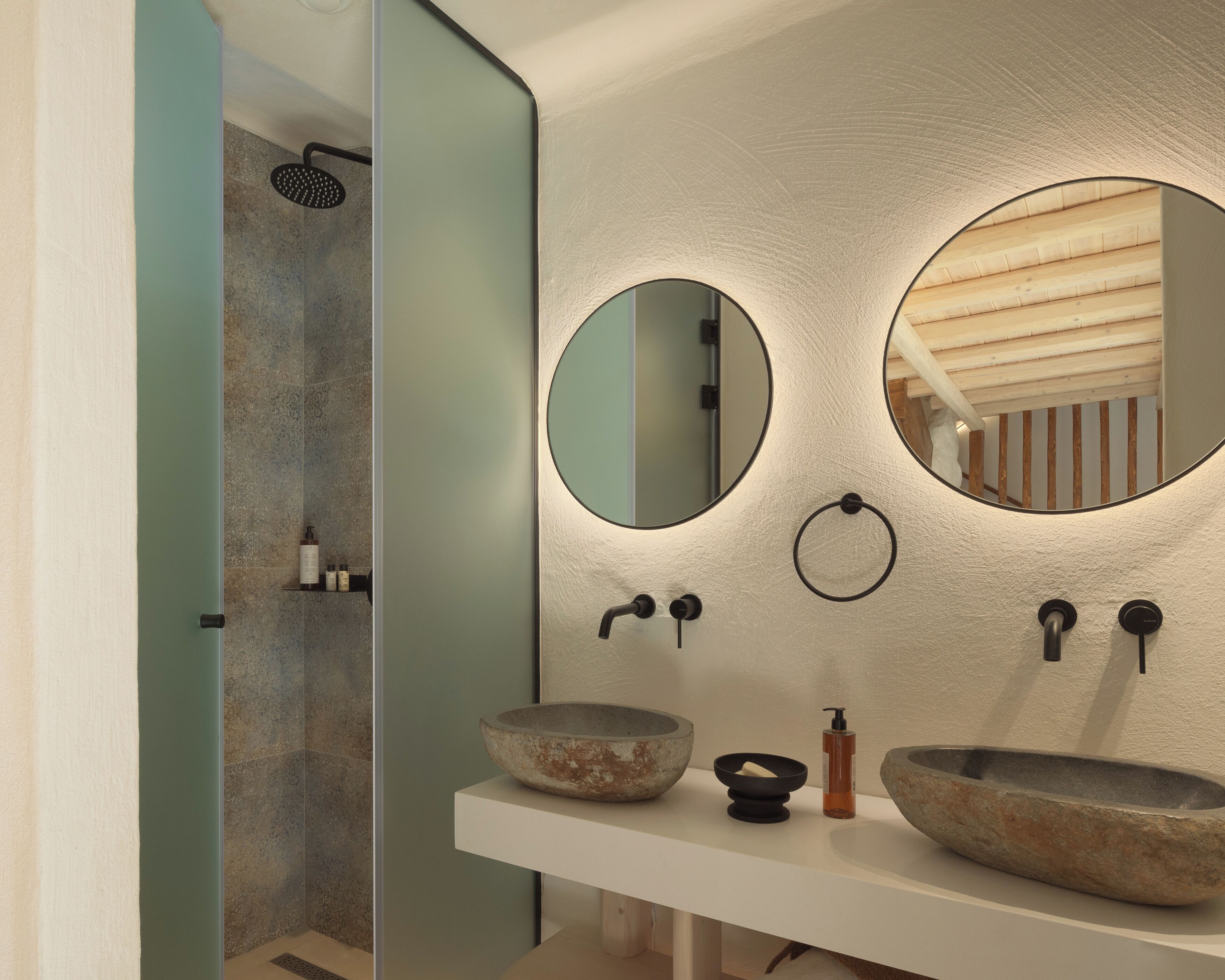 A bathroom with a white vanity, two round mirrors, a stone sink, and a shower area with a glass door.