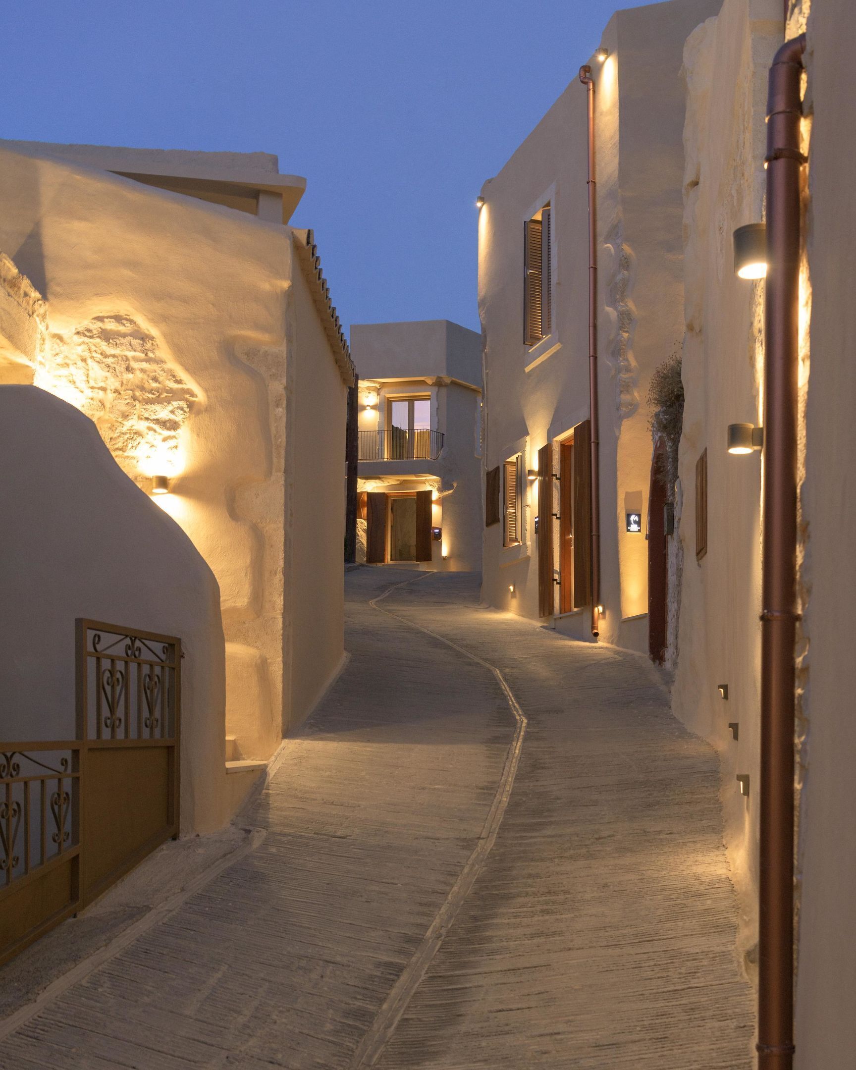 A narrow cobblestone street flanked by buildings with warm lighting, leading to a central open area with a staircase.