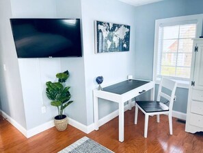 Una habitación con un televisor de pantalla plana montado en la pared, una obra de arte con un mapa mundial, un escritorio blanco con una silla, una planta en maceta y una ventana con persianas.