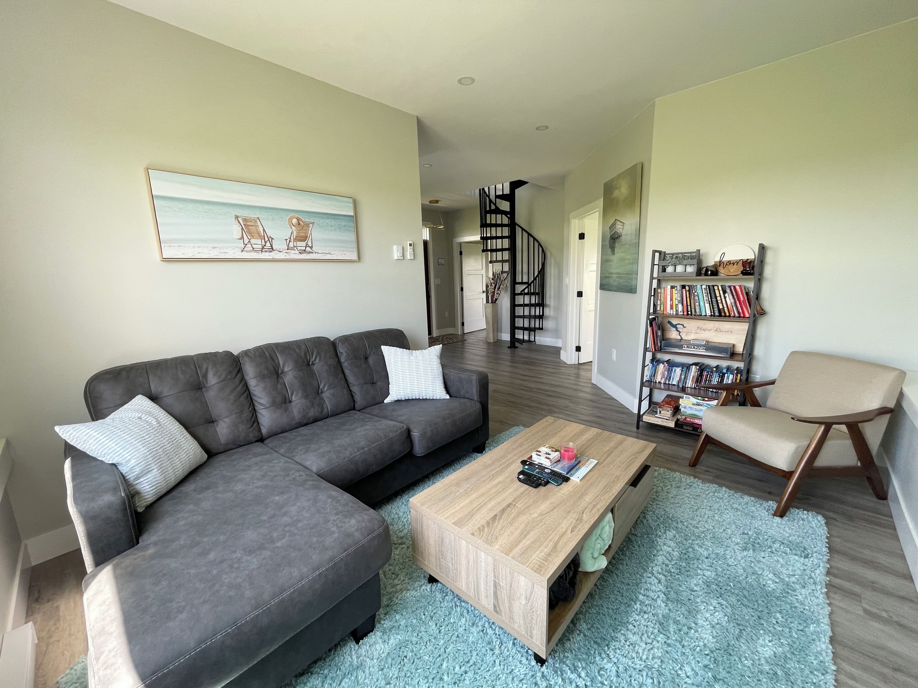 A living room with a grey sectional sofa, a wooden coffee table, a bookshelf, and a spiral staircase in the background.