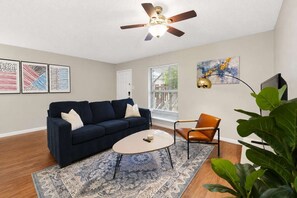 Un salon avec un canapé bleu foncé, une table basse ronde, un tapis à motifs, un ventilateur de plafond et une grande fenêtre avec des stores.