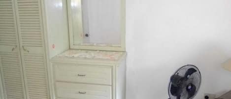 Une chambre à coucher avec un lit, une commode, un miroir et un ventilateur sur pied.