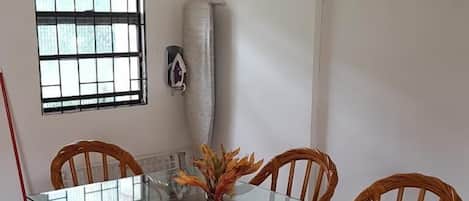 A dining area with a glass-top table surrounded by wicker chairs, a vase with dried flowers, and a window with bars.
