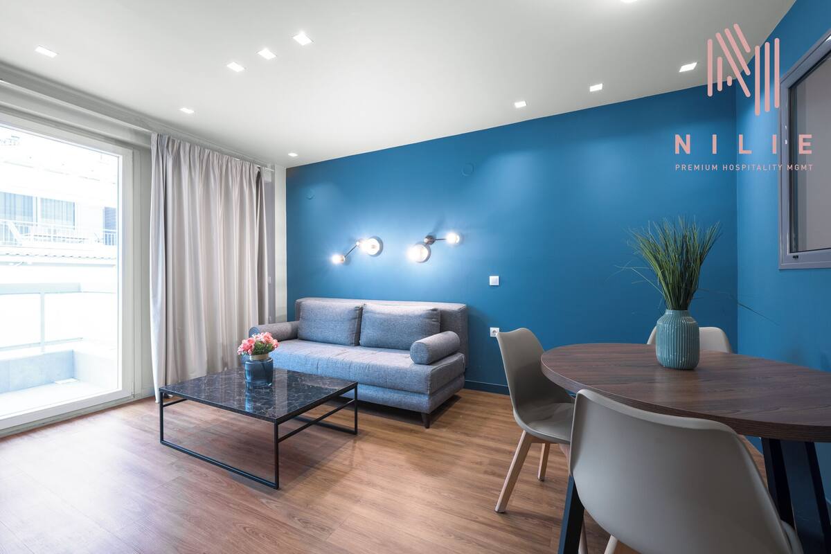 Una sala de estar moderna con un sofá, una mesa de centro y una mesa de comedor con sillas. Hay un ventanal con cortinas, una pared azul decorativa y un jarrón decorativo en la mesa de comedor.
