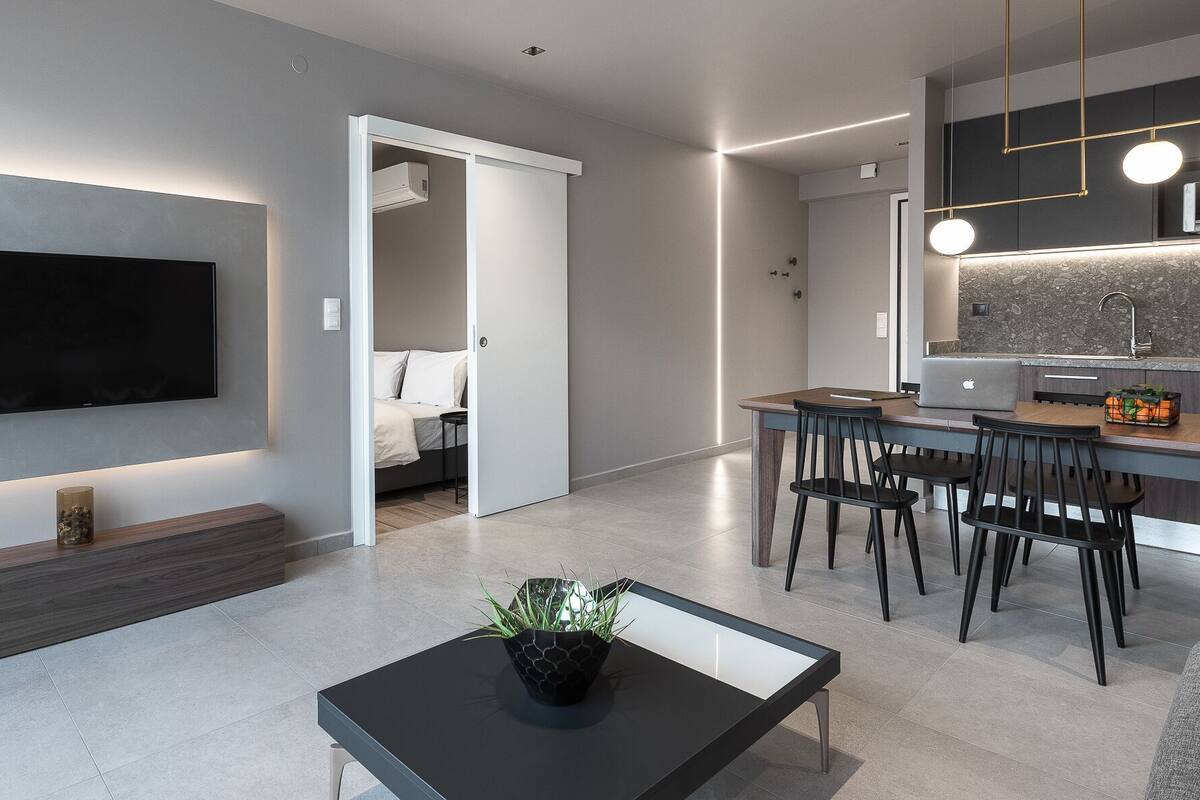 Una sala de estar moderna con un televisor de pantalla plana, una mesa de centro con una planta, y un comedor con sillas y una computadora portátil.