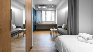 A modern hotel room with a wooden floor, a bed, a sofa, a small table with plants, and a window with curtains.