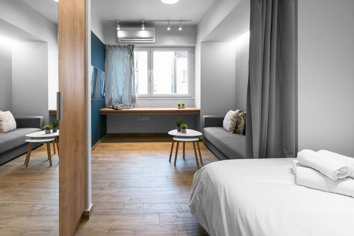 Ein modernes Hotelzimmer mit Holzboden, einem Bett, einer Couch, einem kleinen Tisch mit Pflanzen und einem Fenster mit Vorhängen.