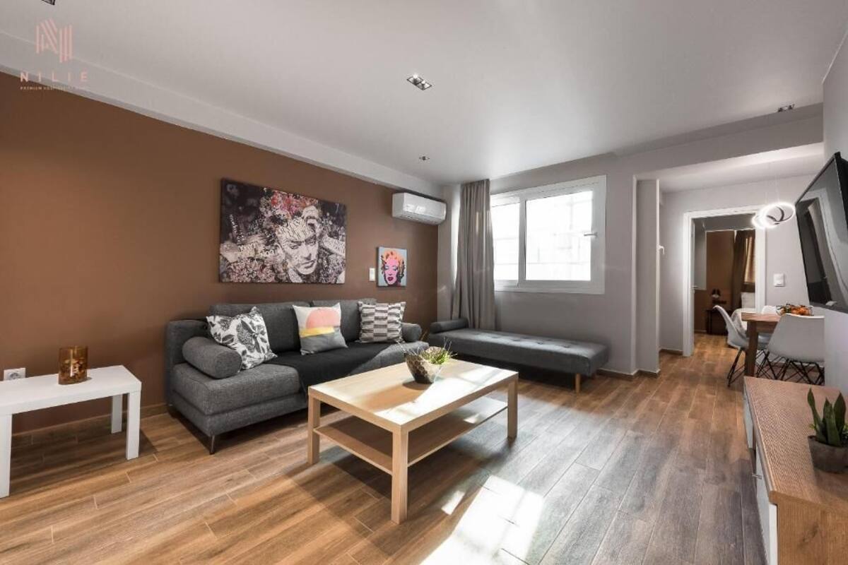 Una sala de estar moderna con un sofá gris, una mesa de centro de madera y una mesita auxiliar blanca. Hay un cuadro en la pared y una ventana con cortinas.