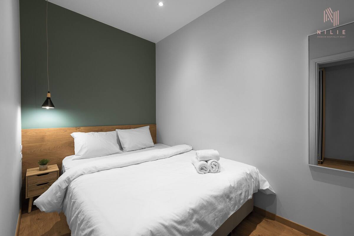 Una cama bien tendida con sábanas blancas, una mesita de noche con una planta y una lámpara colgante.