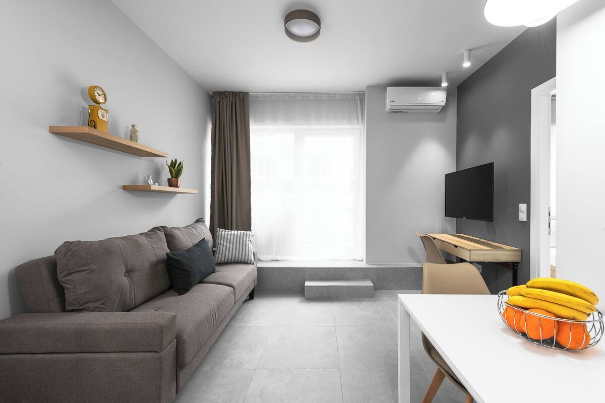 Una sala de estar moderna con un sofá gris, una mesita, un televisor y una ventana con cortinas.