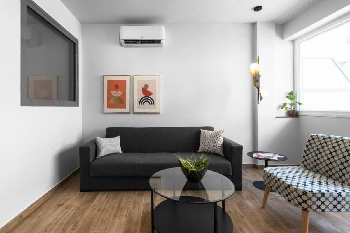 Una sala de estar moderna con un sofá negro, una mesa de centro redonda de vidrio y un sillón estampado. Hay un aire acondicionado instalado en la pared y dos obras de arte enmarcadas sobre el sofá.