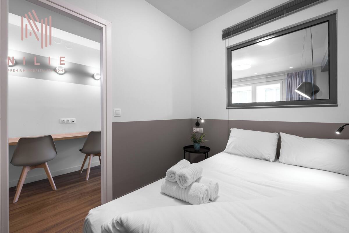 Habitación de hotel con una cama, dos sillas, una mesita de noche con lámpara, una planta en maceta y un espejo grande en la pared.