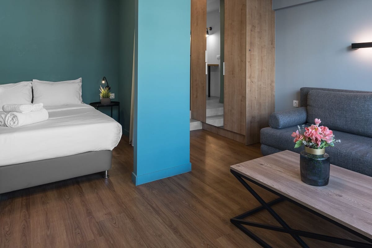 Ein modernes Hotelzimmer mit Bett, einer Couch, einem Couchtisch mit Blumenvase und einem Flur, der in einen weiteren Raum führt.