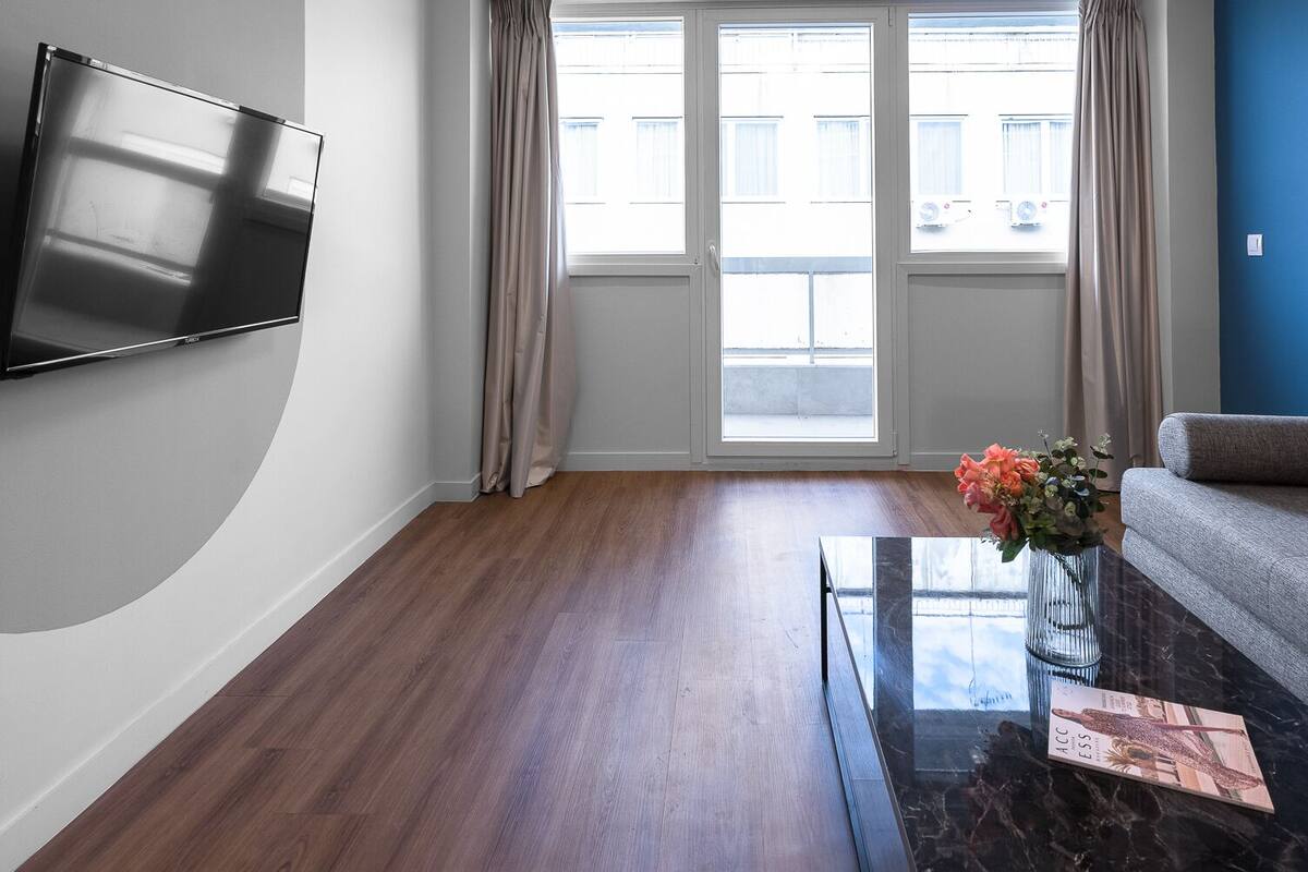 Una sala de estar moderna con un televisor de pantalla plana montado en la pared, un sofá gris, una mesa de centro con un florero y un ventanal con cortinas.