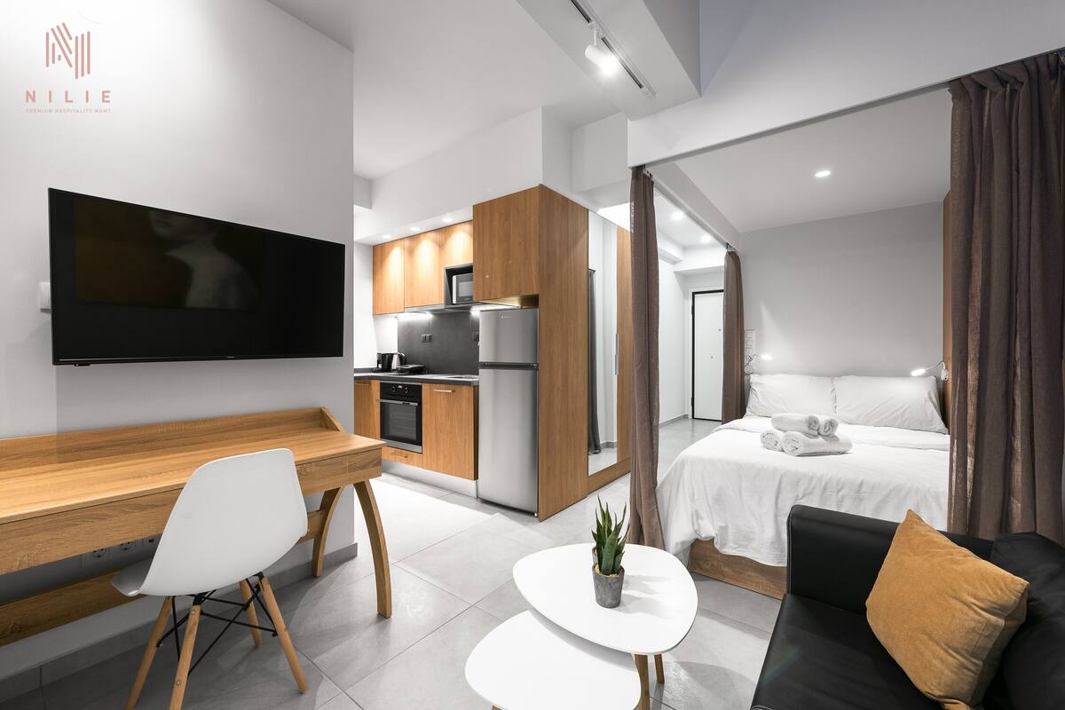 Ein modernes Hotelzimmer mit einem Holzbüro, einem weißen Stuhl, einem kleinen weißen Tisch mit einer Pflanze, einem schwarzen Sofa mit einem braunen Kissen, einem Bett mit weißen Bettwäsche und einer Küchenzeile mit Kühlschrank und Mikrowelle.