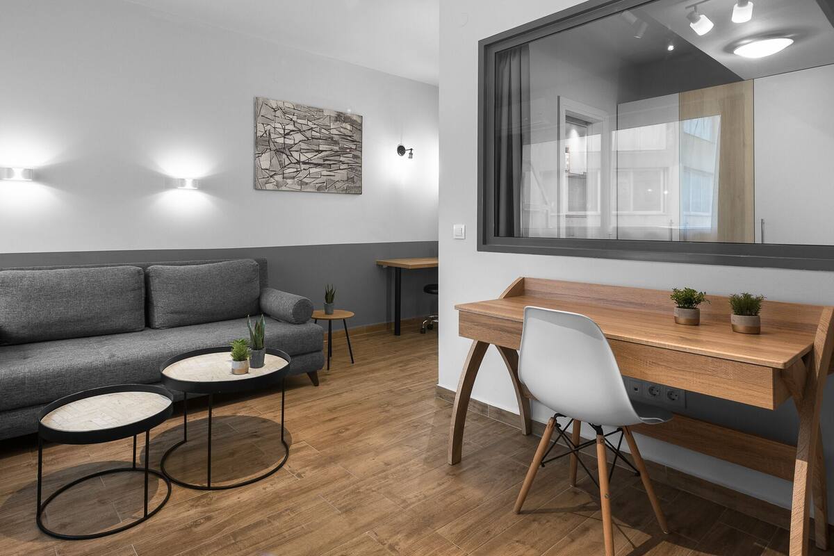 Una habitación moderna con un sofá gris, dos mesas redondas, un escritorio de madera con una silla y un espejo grande.