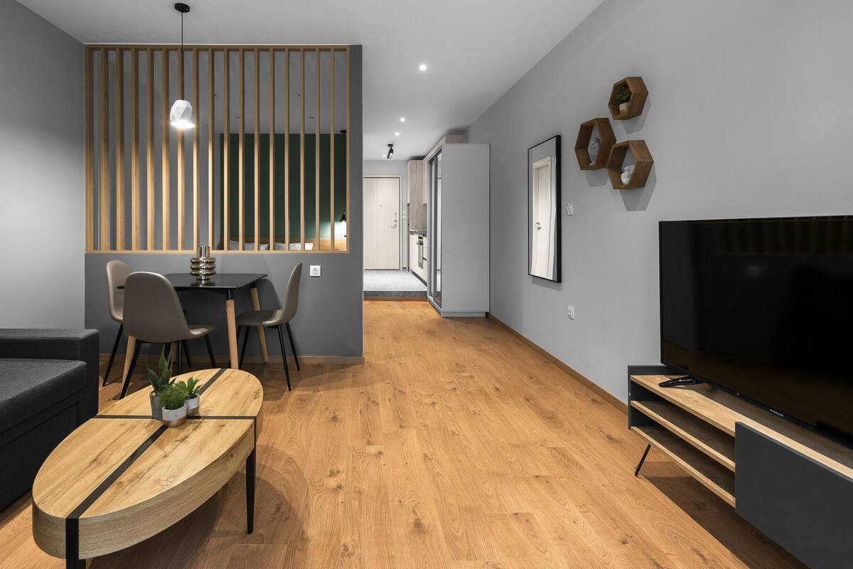 Una sala moderna con un sofá, una mesa de centro y un televisor. Hay una zona de comedor con sillas y una mesa, y una cocina al fondo.