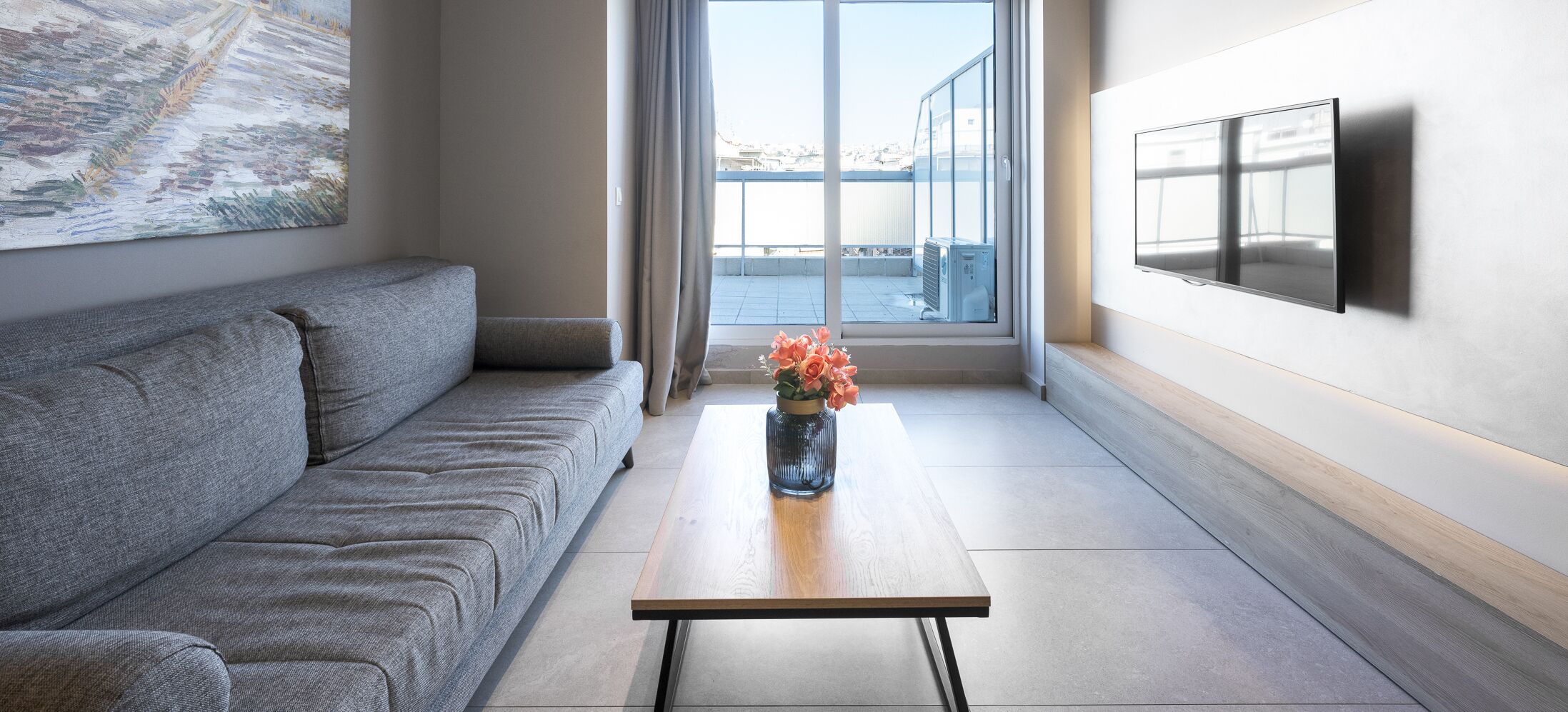 Een moderne woonkamer met een grijze bank, een salontafel met een bloevas en een aan de muur bevestigde televisie.