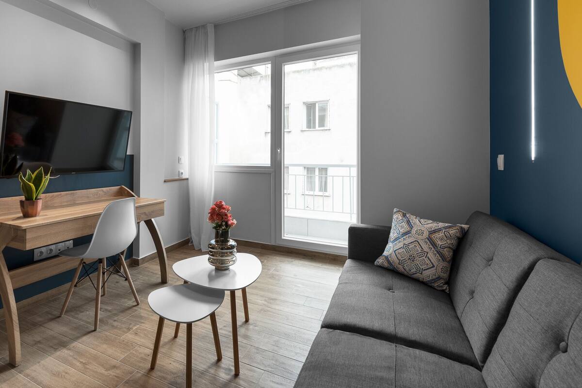 Ein modernes Wohnzimmer mit Flachbildfernseher, einem Holztisch, einem grauen Sofa und einem kleinen runden Tisch mit einer Blumenvase.