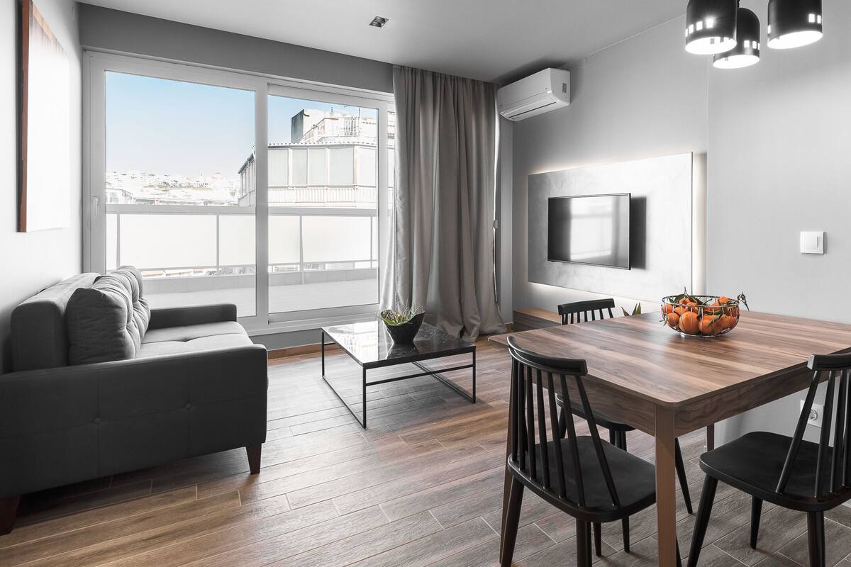 Una sala de estar moderna con un sofá, una mesa de comedor con sillas, un televisor montado en la pared y un ventanal con cortinas.