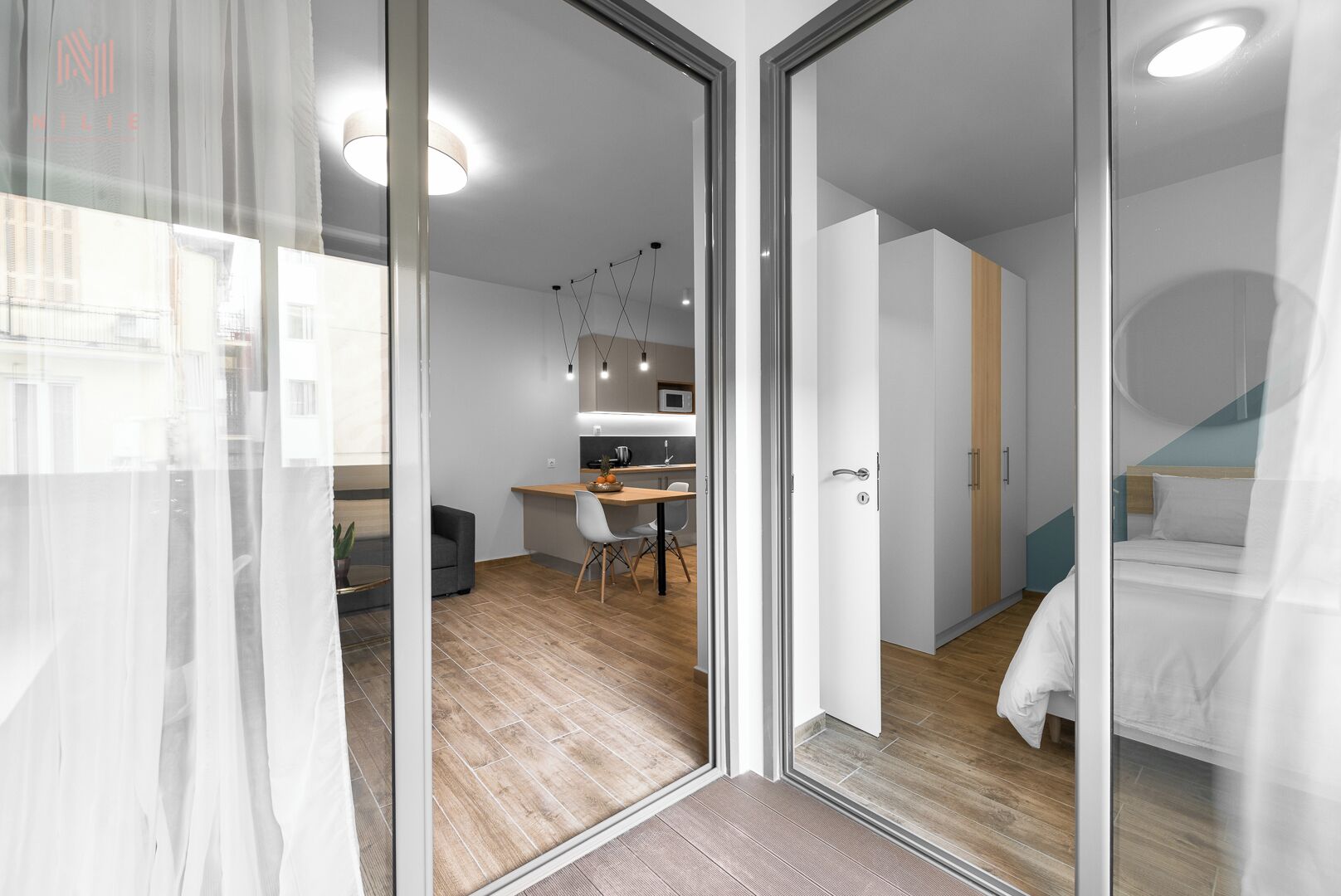 Una habitación moderna y luminosa, con piso de madera, una cama, un escritorio con silla, y un área de cocina visible a través de una puerta de vidrio.