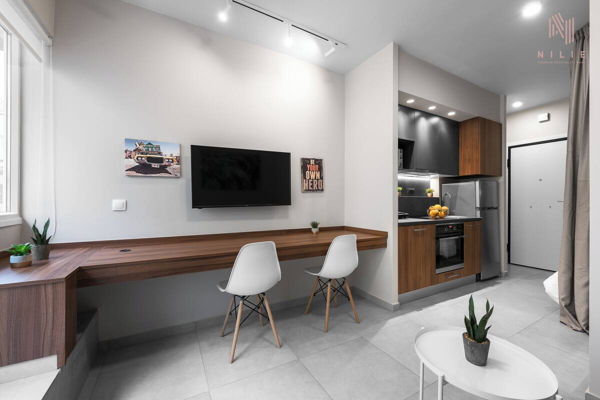Eine moderne Küche mit Holzausziehtisch, zwei weißen Stühlen, einem an der Wand befestigten Fernseher und einem kleinen Topfpflanzen auf der Arbeitsfläche.