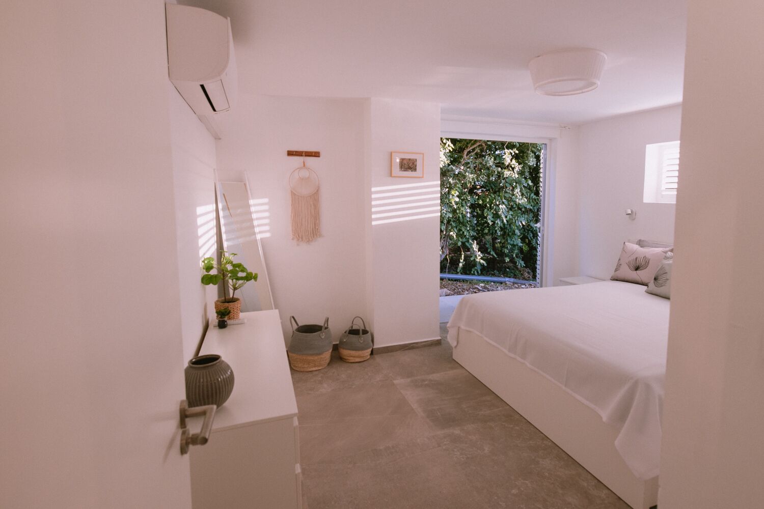 Une chambre à coucher avec un lit, une table de chevet, un miroir, une fenêtre donnant sur un paysage verdoyant et une petite plante sur la table.