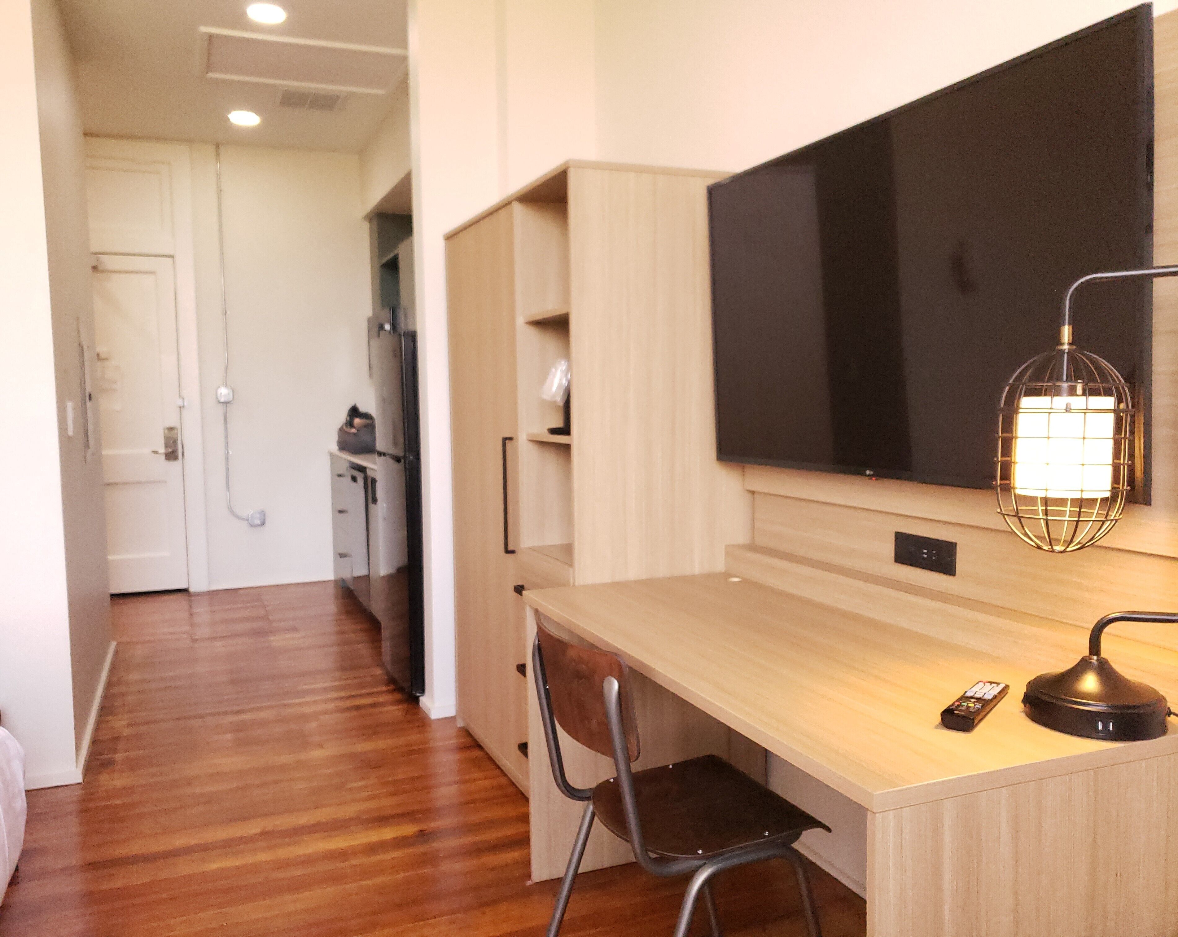 Una habitación de hotel moderna con un escritorio de madera, una silla, un televisor de pantalla plana, una lámpara y un control remoto.