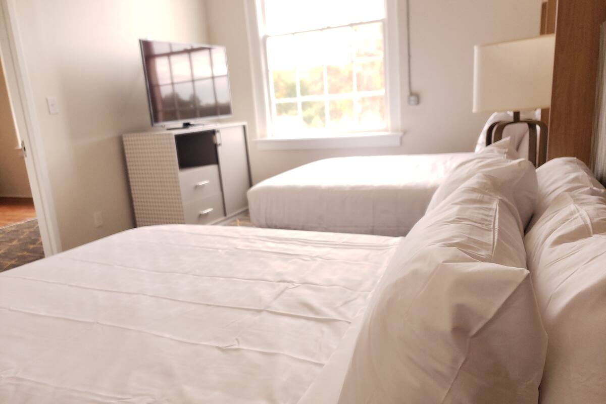 Quarto de hotel com duas camas, uma televisão em uma cômoda e janela com vista para árvores.