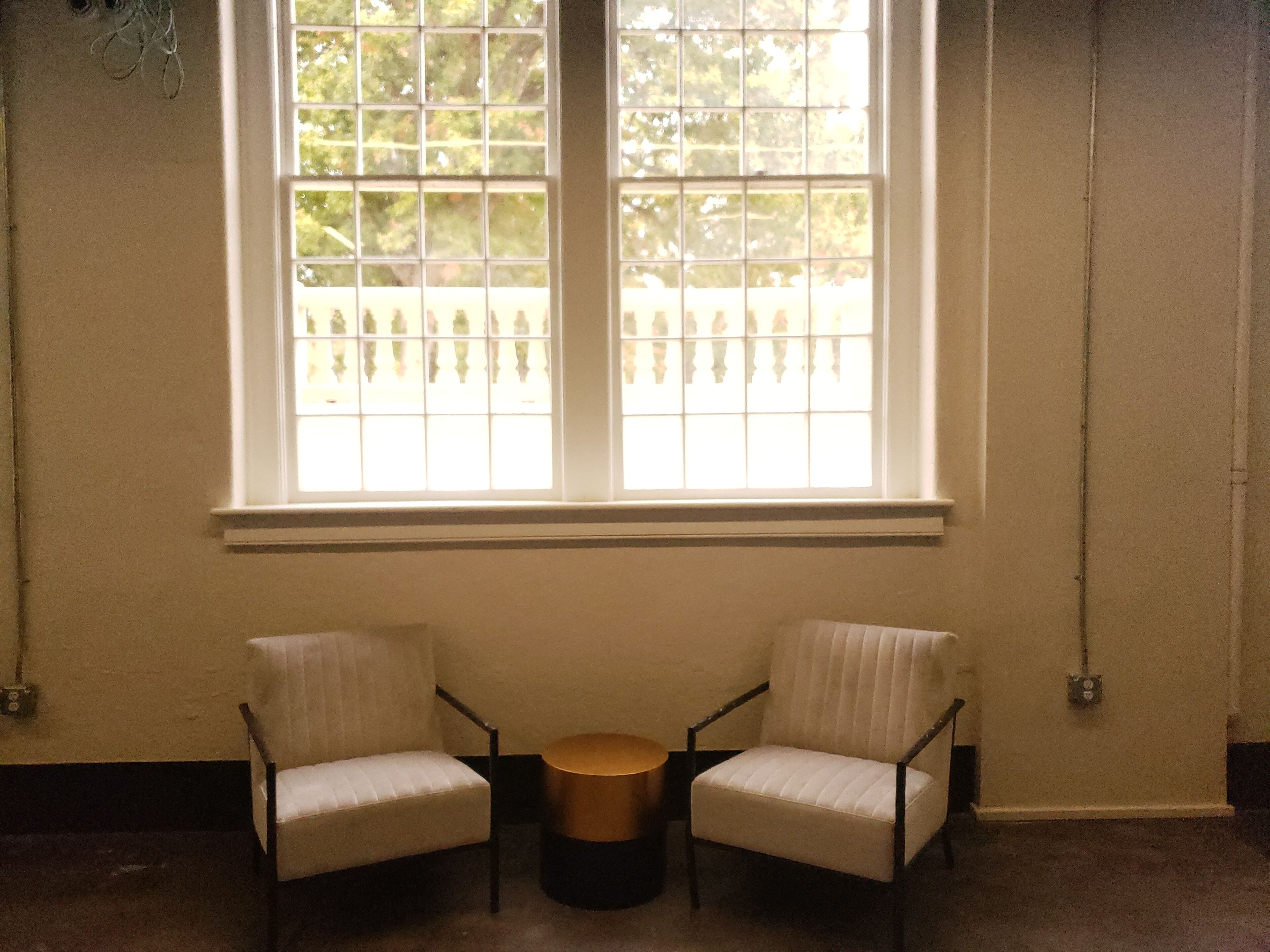 A room with two chairs and a small round table, a large window with a view of a fence and trees, and a plain wall with a switchboard.