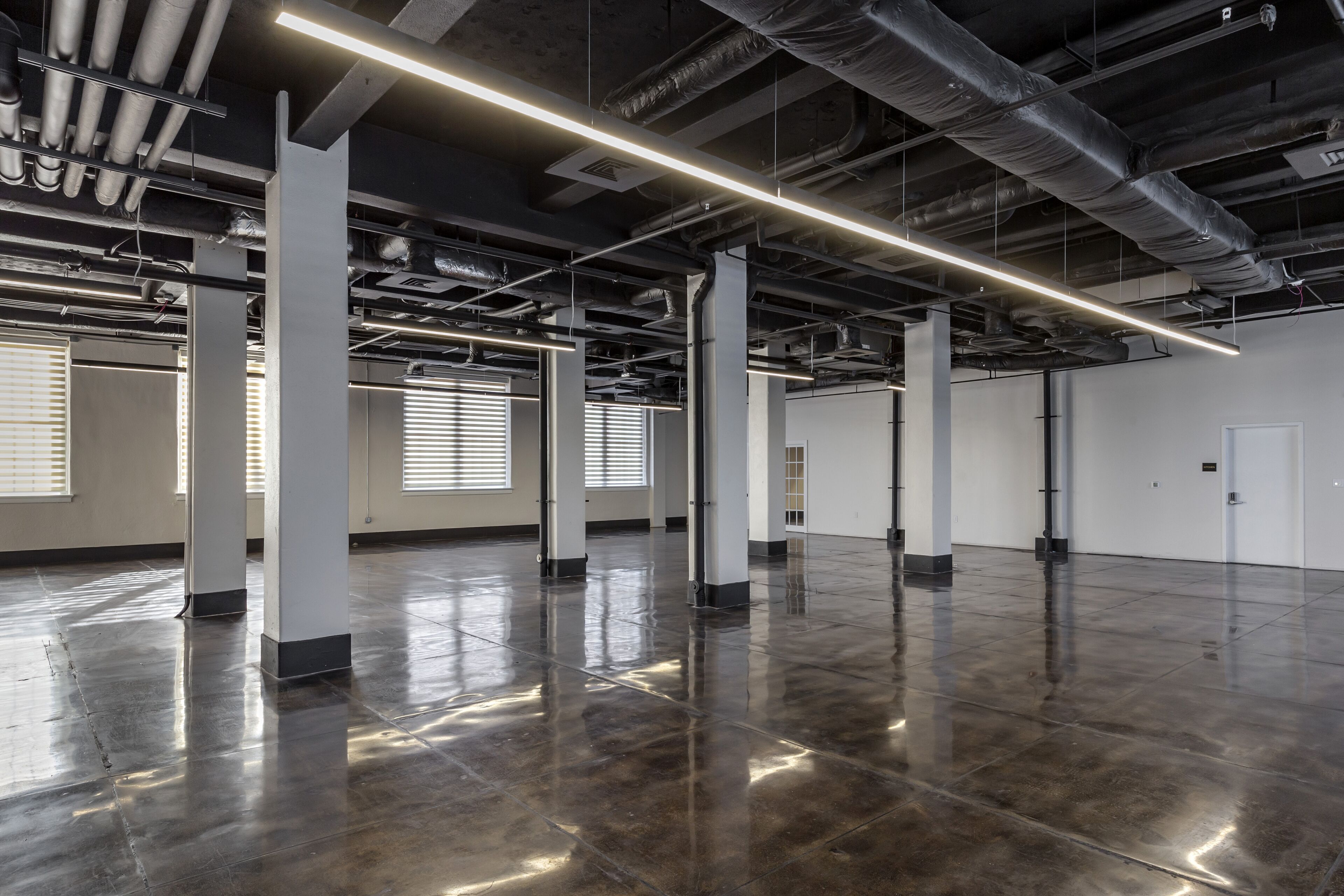 A spacious, industrial-style interior with exposed ductwork, polished concrete floors, and large windows with blinds.