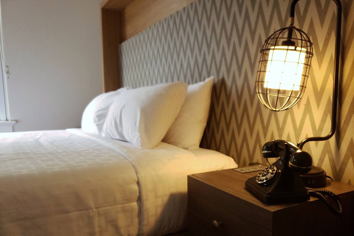 Uma cama bem arrumada com roupa de cama branca, um telefone antigo em uma mesa de cabeceira e uma parede com padrão em zigue-zague.