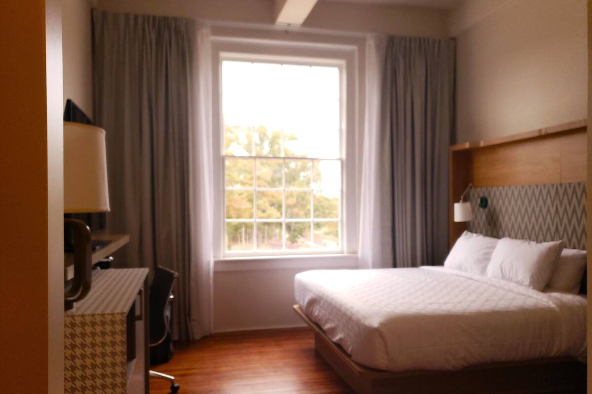 Quarto de hotel com uma cama grande, uma escrivaninha com abajur e uma janela com cortinas.