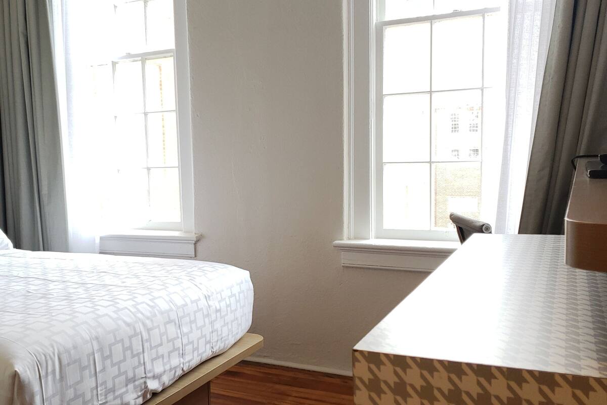 Quarto de hotel com uma cama, uma escrivaninha e duas janelas.