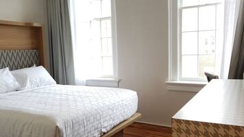 A hotel room with a bed, a desk, and two windows.