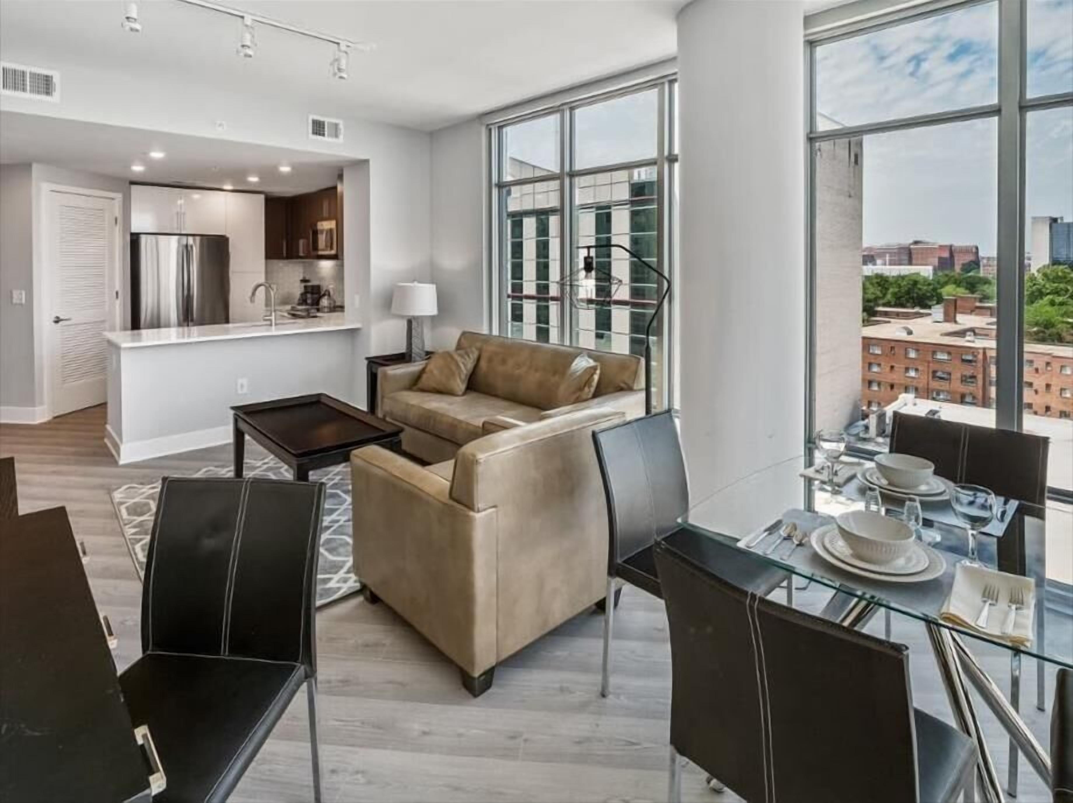A modern living room with a sofa, dining table, and chairs, a kitchen area, and large windows offering a city view.