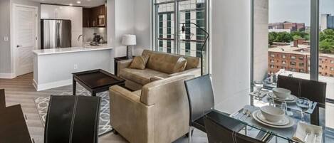 A modern living room with a sofa, dining table, and chairs, a kitchen area, and large windows offering a city view.