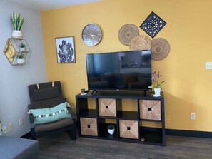 A living room with a TV on a black stand, a sofa, a wall clock, decorative wall art, and a small plant. - Lover's Hideaway (Anderson)