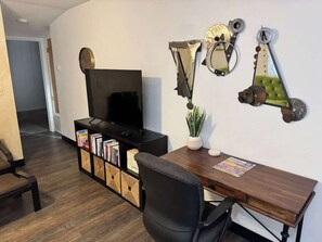 A modern living room with a flat-screen TV, a wooden dining table, a black office chair, a bookshelf, and decorative wall art.