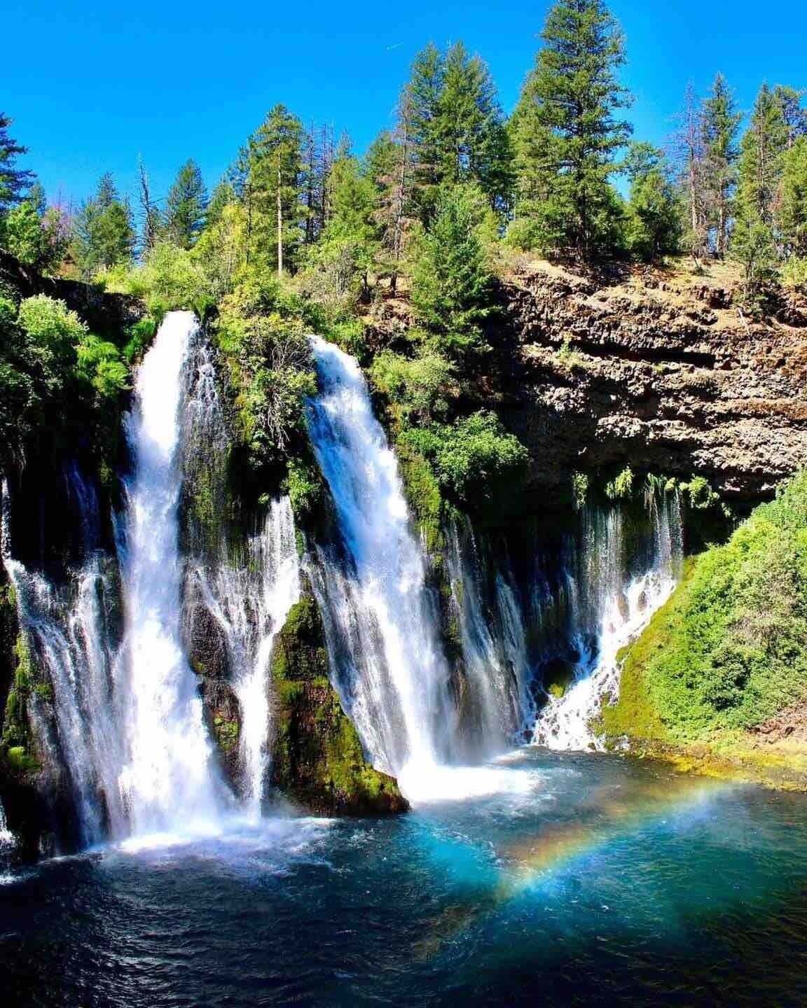 Uma cascata com vários riachos descendo em direção a um lago, rodeada por vegetação exuberante e árvores.