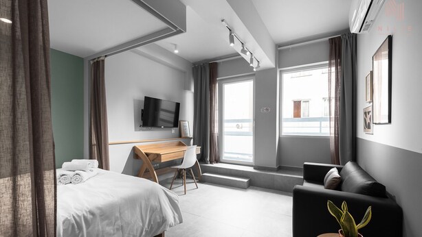 Une chambre d’hôtel moderne équipée d’un lit, d’un bureau et d’un coin salon, avec une télévision, de grandes fenêtres et une plante en pot.