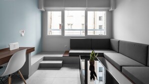Une chambre d’hôtel moderne et minimaliste, avec un canapé gris, un bureau en bois et une grande fenêtre donnant sur la ville.