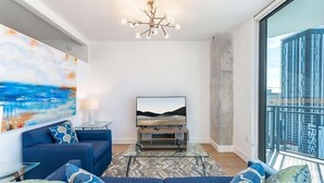 Una sala de estar moderna con un televisor grande, sofás azules, una mesa de centro de vidrio y vistas a la ciudad a través de ventanas amplias.