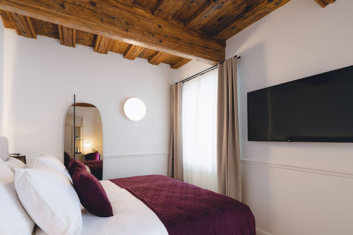 A hotel room with a wooden ceiling, a bed with white and maroon bedding, a round mirror, a flat-screen TV, and a window with curtains.
